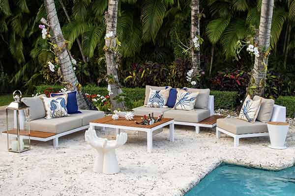Luxurious outdoor lounging area with contemporary beige sofas adorned with navy and white nautical-themed pillows, a wooden coffee table, and unique white seating, set against a backdrop of tropical foliage and a poolside setting.