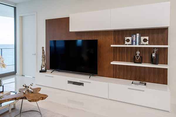 Elegant TV wall unit with dark wood backdrop, white cabinets, floating shelves with decor, and seaside view in a bright room 