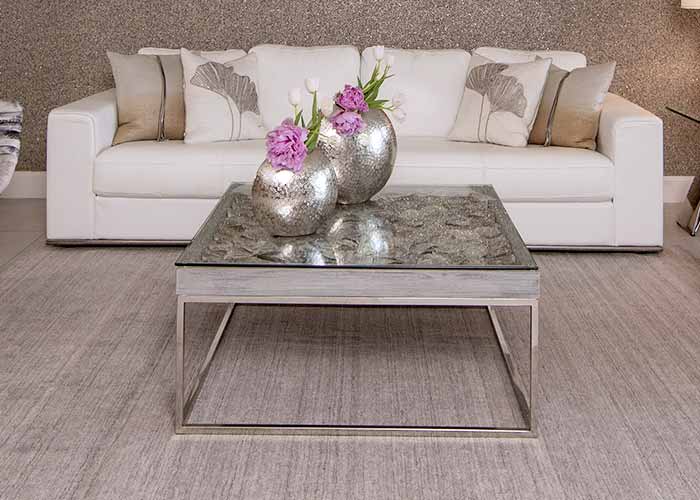 The Infinity coffee table, a modern piece with a reflective glass top and unique base, central in a living room with a white sectional sofa.