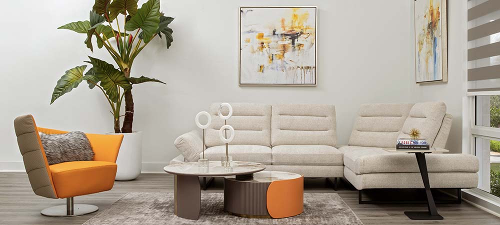 Modern living room showcasing a white leather couch with gold pillows, contemporary tan armchairs, a clear glass center table, abstract wall art, a decorative ceramic lamp, and a multi-colored area rug.