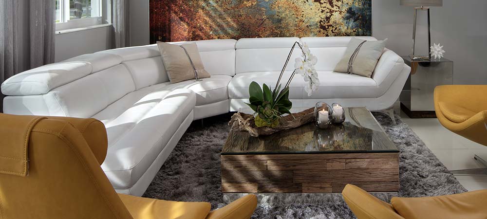 Chic living space featuring a curvaceous white leather sectional sofa, rustic wooden coffee table with greenery centerpiece, golden yellow accent chairs, plush gray rug, and an abstract canvas painting, complemented by soft natural light and elegant drapery.