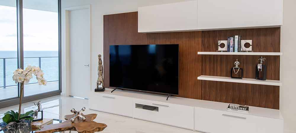 Elegant TV wall unit with dark wood backdrop, white cabinets, floating shelves with decor, and seaside view in a bright room
