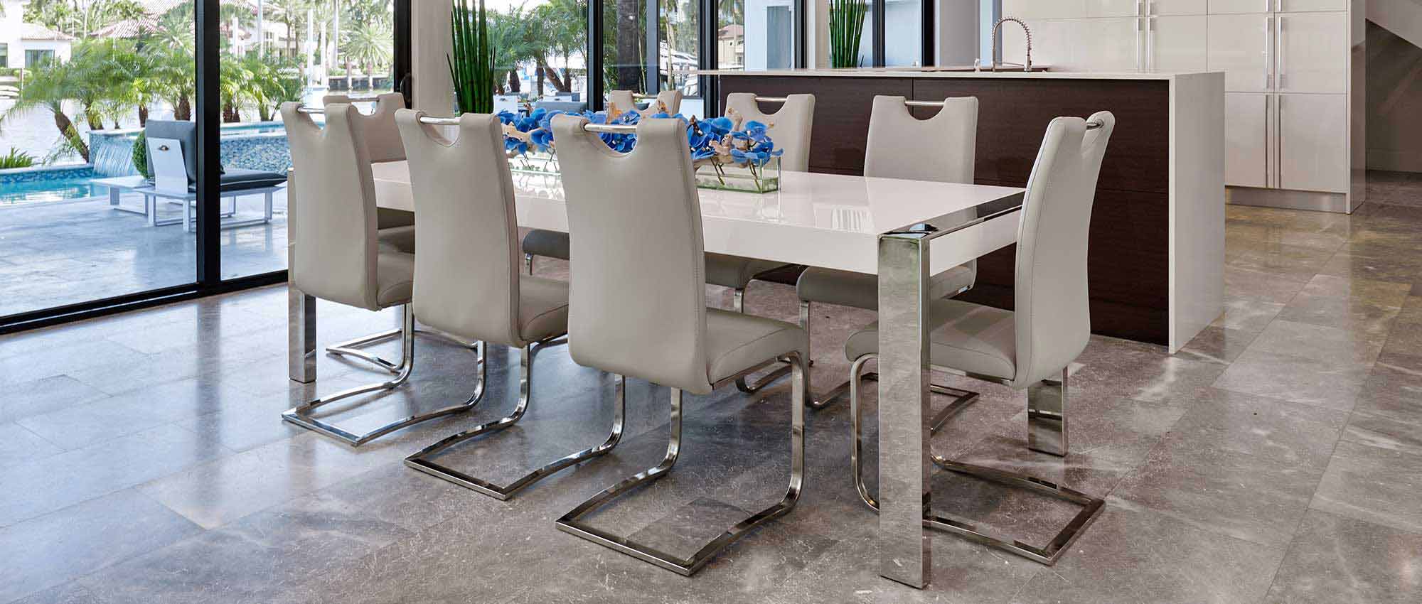 A modern dining room with a white lacquer dining table, grey chairs, and a blue orchid floral table centerpiece. TThere is a large window that lets in natural light.
