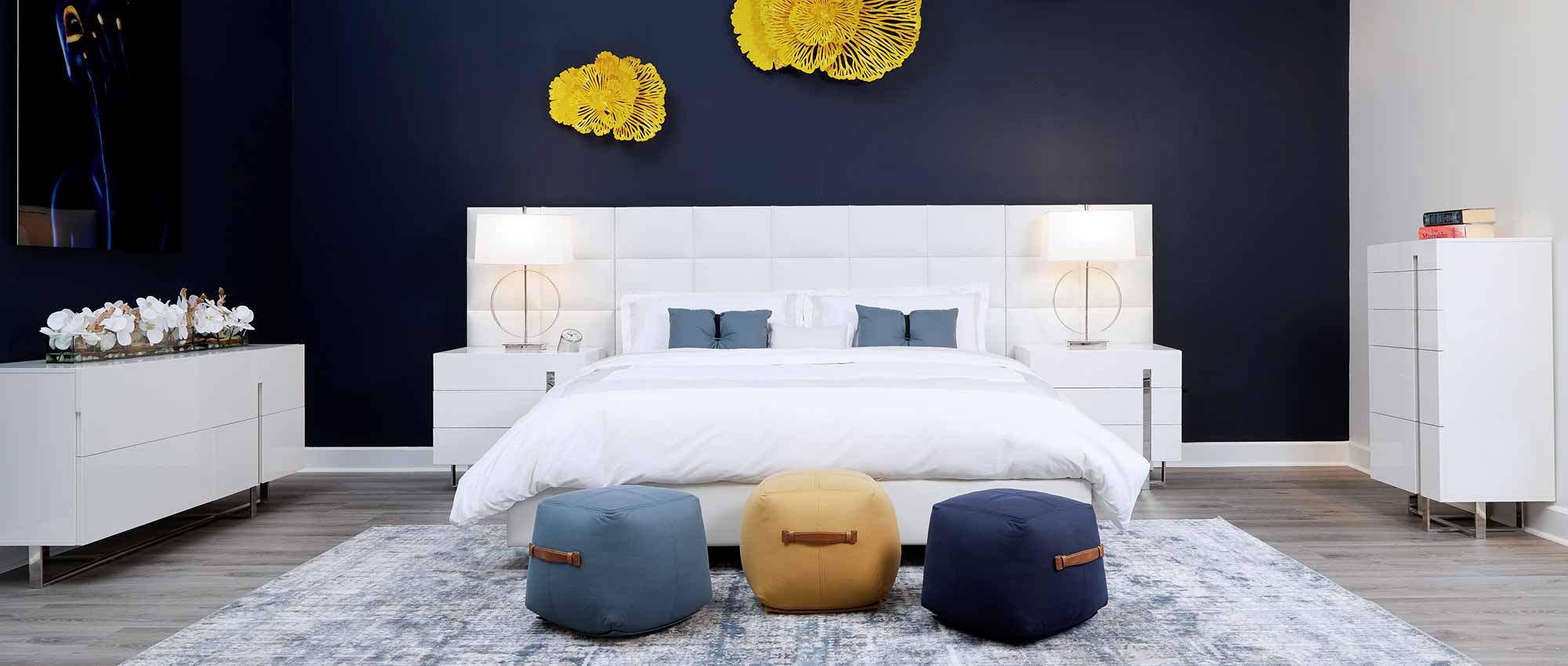  A modern bedroom with a white eco-leather bed, a white nightstand, and a white dresser. The walls are decorated with modern art. The bed is covered in a white duvet, and there are two pillows in shades of blue. The nightstand has a modern lamp, and a clock. The overall style of the room is clean and modern.