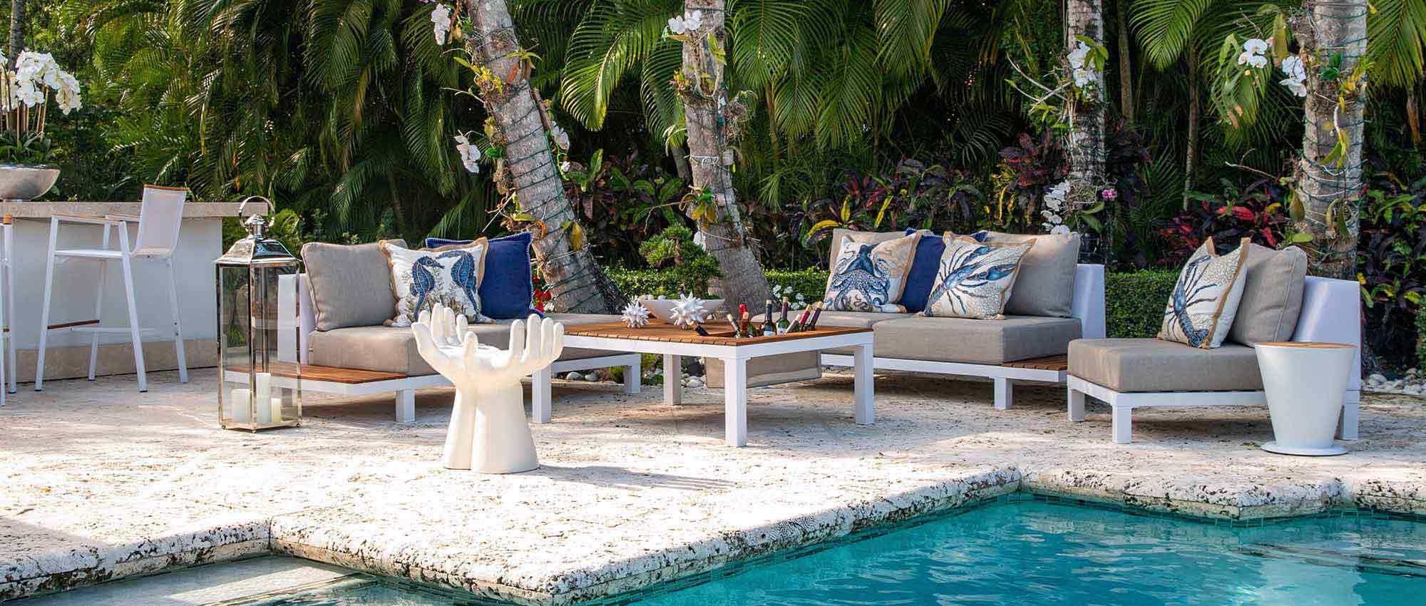 A modern outdoor lounging furniture set with a sofa, two gray chairs, and a teak top coffee table. The furniture is made of weather-resistant materials, and it is designed to be comfortable and stylish. The furniture is placed on a patio, and there is a view of a pool and lush landscaping.