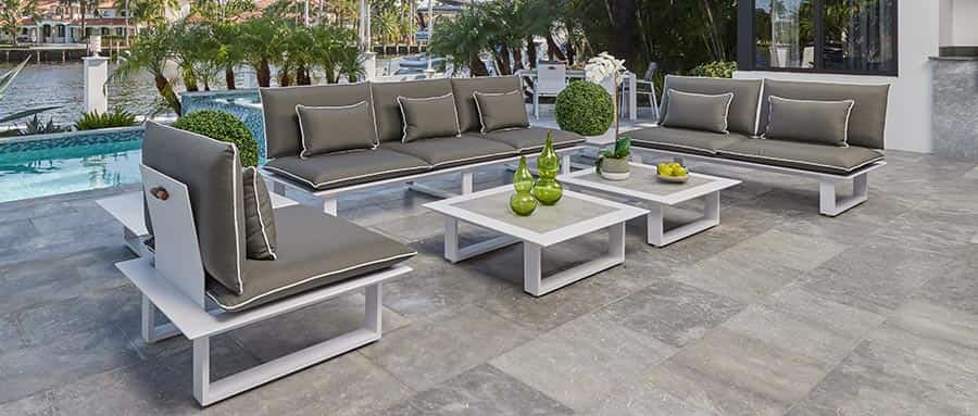A modern Outdoor area of a house with a gray outdoor sofa, a white outdoor coffee table with ceramic top that has a white orchid floral and a modern accessory as decoration.
